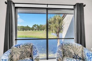 Ein Zimmer mit großem Fenster, zwei gemusterten Sesseln und Blick auf eine Grünfläche mit Bäumen und einem Gewässer.