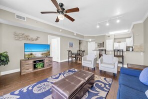 A spacious living room with a ceiling fan, a flat-screen TV, a wooden entertainment center, and a dining area with chairs. - 🌴Tidewater 2112-Gulf Front-Heated Pool-Hot Tub-Gym and MORE🌴 (Panama City Beach)