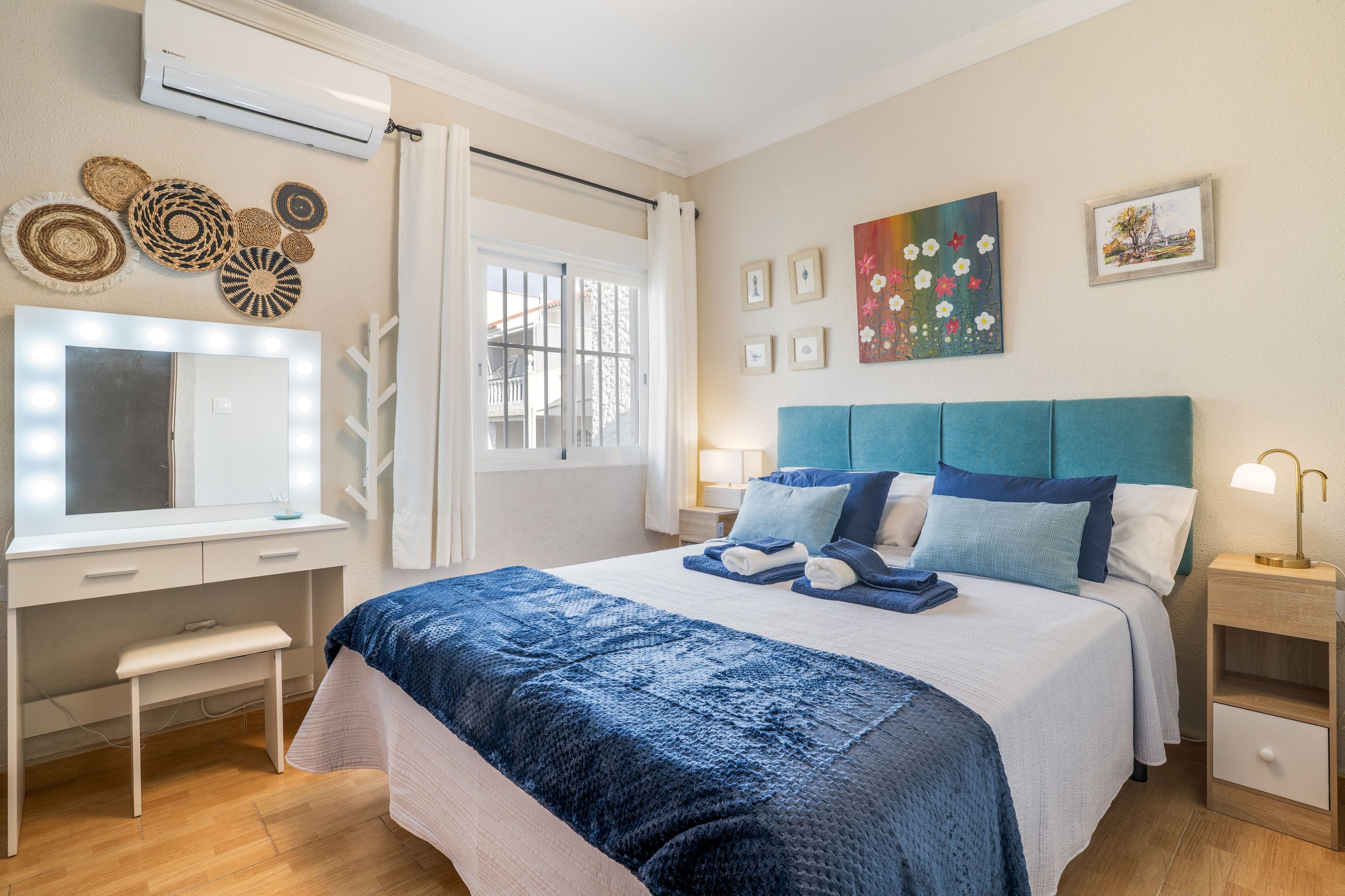 A hotel room with a bed, bedside tables, a desk with a mirror, wall art, and an air conditioning unit.