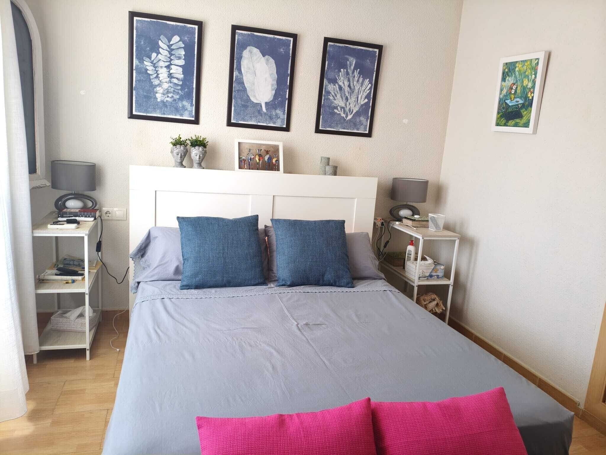 Un dormitorio con una cama, dos mesitas de noche, un escritorio y una silla. Hay tres obras de arte enmarcadas en la pared.