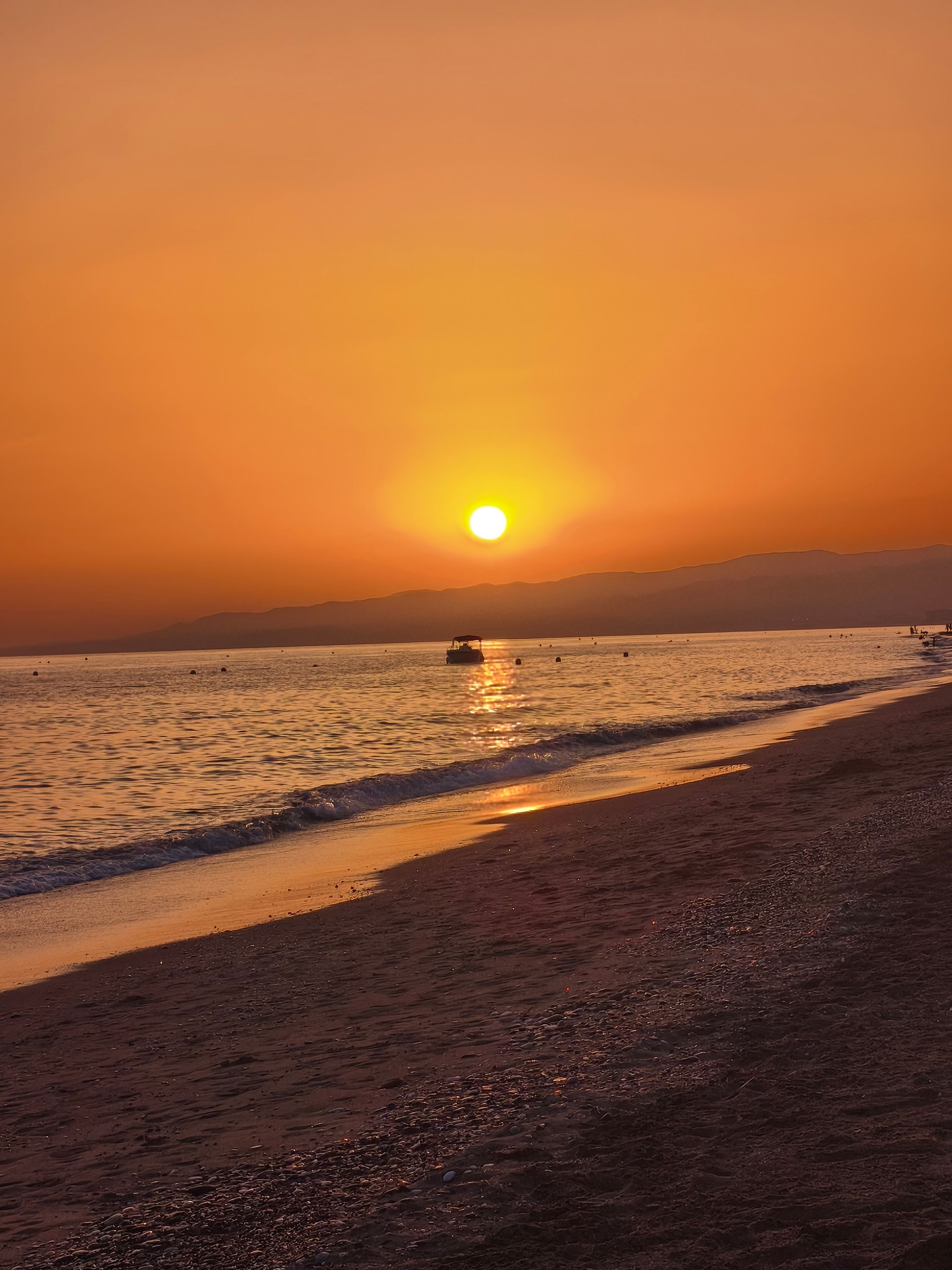 Escena de playa con atardecer, agua tranquila y montañas a lo lejos.