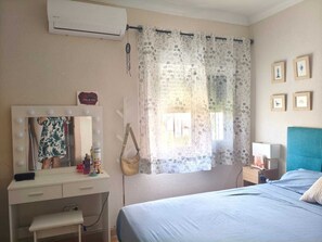 Ein Schlafzimmer mit einem Bett, einem Frisiertisch mit Spiegel und Beleuchtung, einem Fenster mit Vorhängen und Bildern an der Wand.