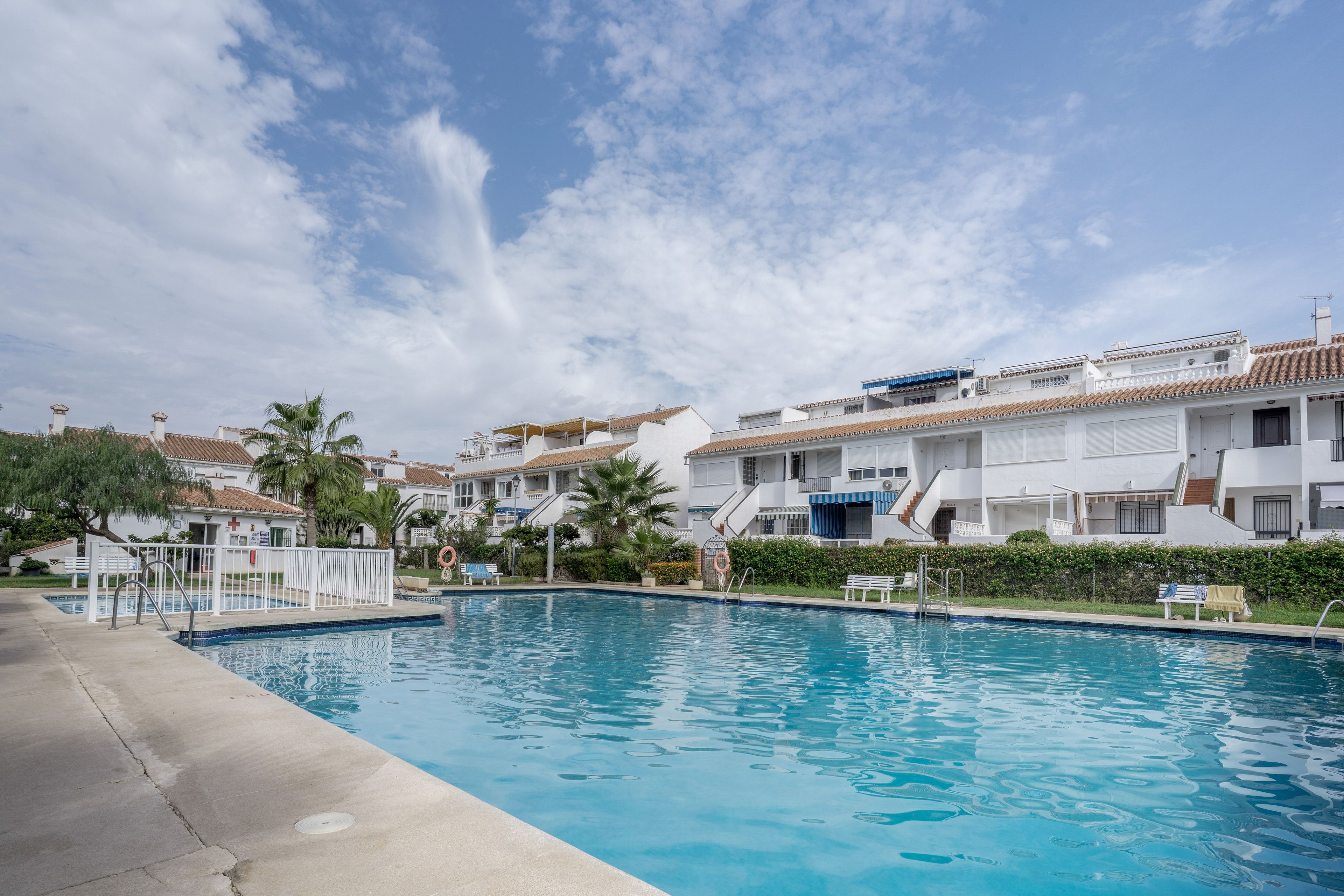 Une piscine, avec un ciel bleu azur au-dessus, entourée de bâtiments blancs aux toits recouverts de tuiles rouges.
