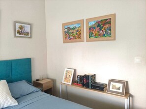 A bedroom with a bed, a nightstand, a bedside table with books and framed pictures, and two wall-mounted paintings.