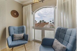 Quarto com duas poltronas azuis, vista para prédios através de uma janela com cortinas brancas e um objeto decorativo na parede.
