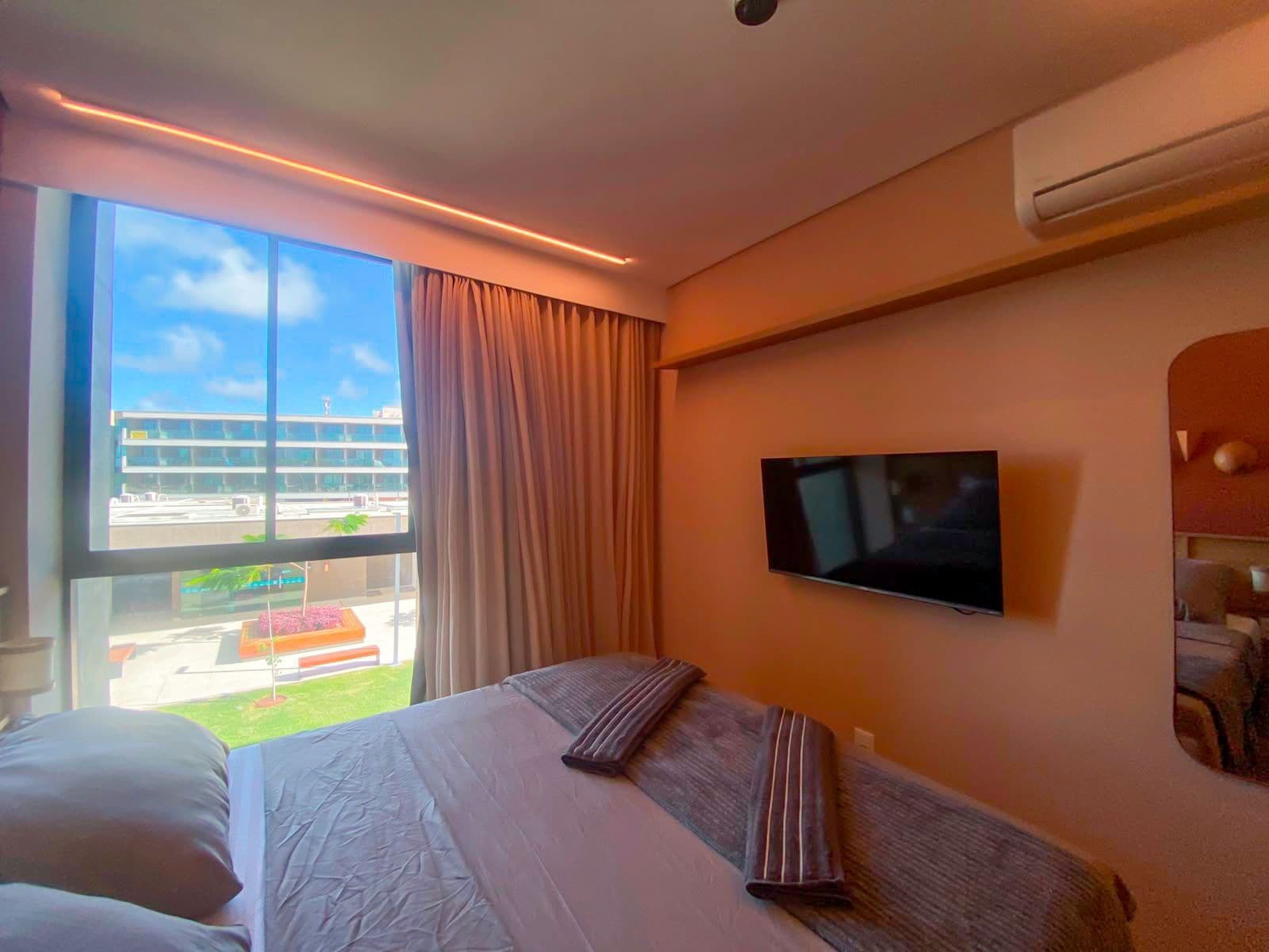 Quarto de hotel com cama, televisão, janela com cortinas e ar condicionado.