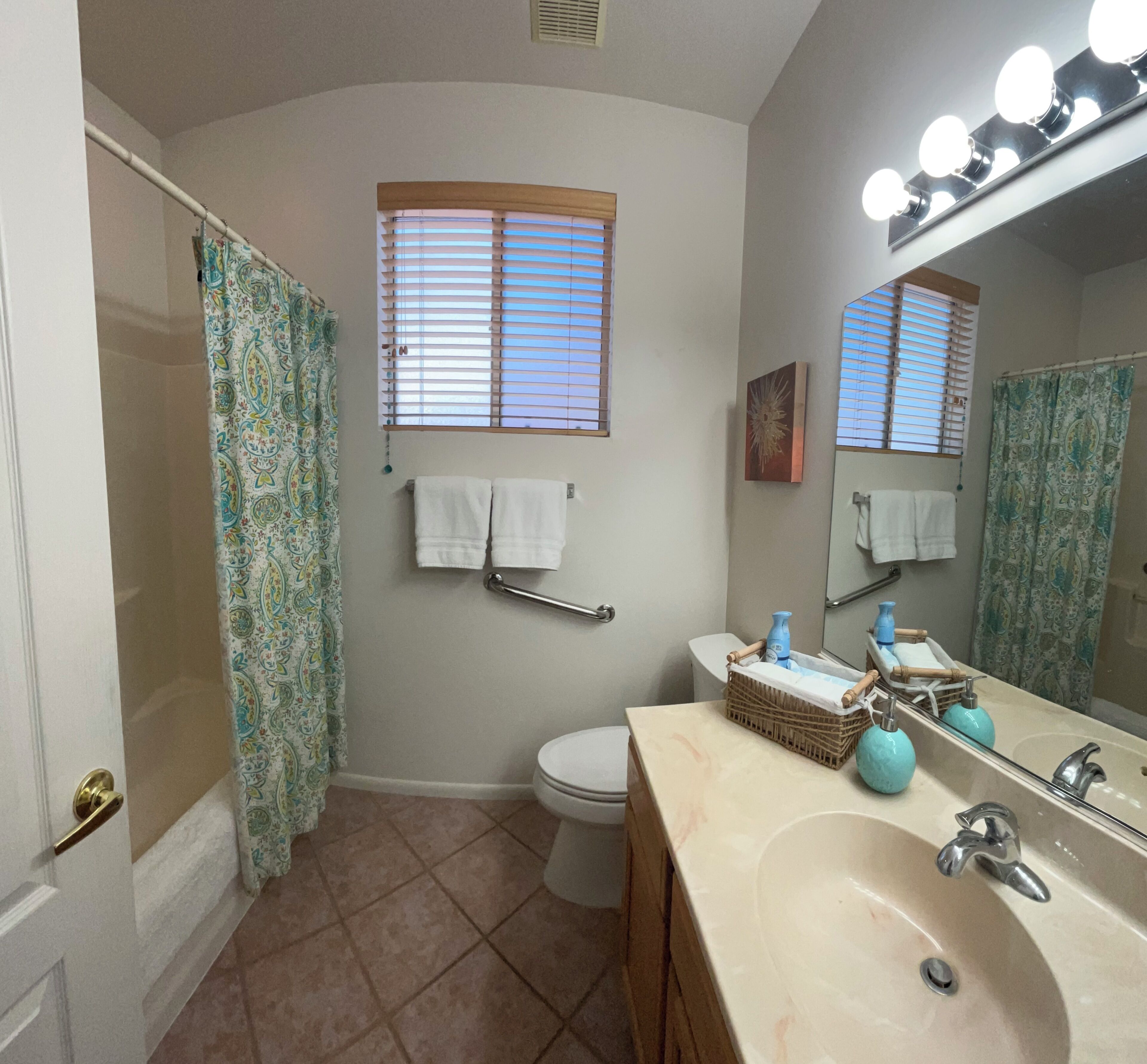 A bathroom with a toilet, sink, and shower area. There are towels hanging on the wall, a basket on the sink, and a mirror with lights above.