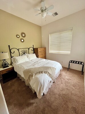 Quarto com cama, mesa de cabeceira, cômoda de madeira, ventilador de teto, janela com persianas e um pequeno banco.