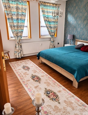 A bedroom with a bed, curtains, a nightstand, a lamp, and a floral-patterned rug. - Apartment 'Sollingnest' with Mountain View, Shared Terrace and Wi-Fi (Uslar)