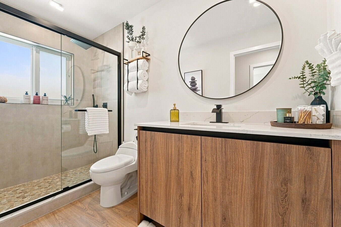 Une salle de bain moderne avec une douche à parois de verre, des toilettes, un meuble-lavabo en bois et un miroir rond.