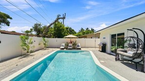Une piscine rectangulaire bordée de blanc, entourée d’une clôture blanche, et une chaise suspendue avec des coussins.