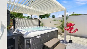Un bain à remous sous une pergola blanche, entouré d’une clôture blanche, avec un banc et un pot de fleurs à proximité.