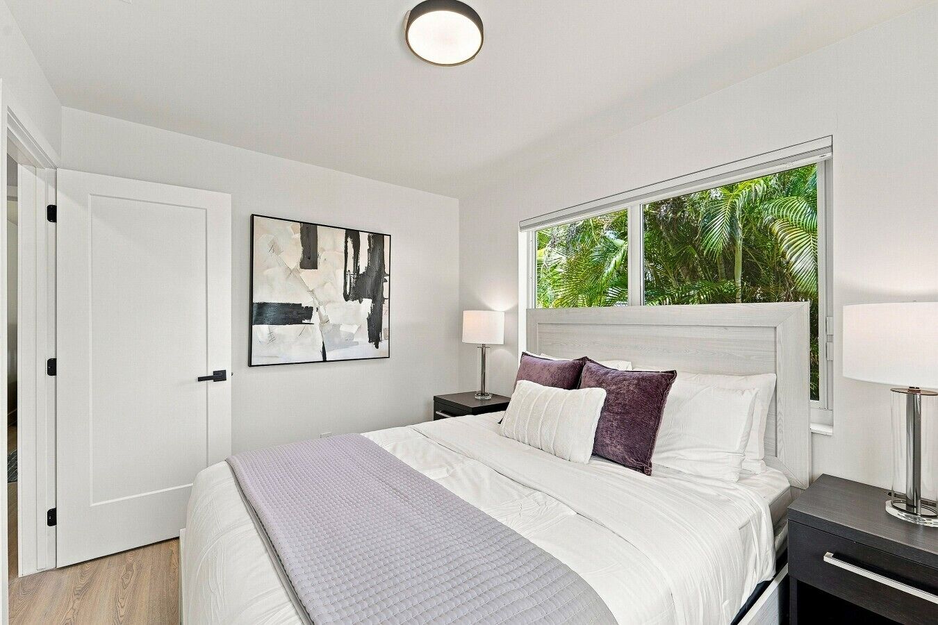 A bedroom with a bed, a nightstand, a lamp, a door, and a window with a view of greenery.