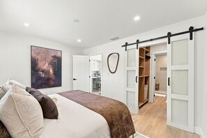 A bedroom with a bed, a large abstract painting, a wardrobe with sliding doors, and a bathroom visible through an open door. - Spa Hot Tub | Mins to Beach | 2 Kings | Brand New! (Pompano Beach)