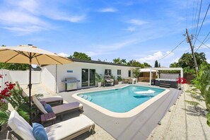 A residential backyard with a swimming pool, lounge chairs, a barbecue grill, and a covered patio area. - Spa Hot Tub | Mins to Beach | 2 Kings | Brand New! (Pompano Beach)