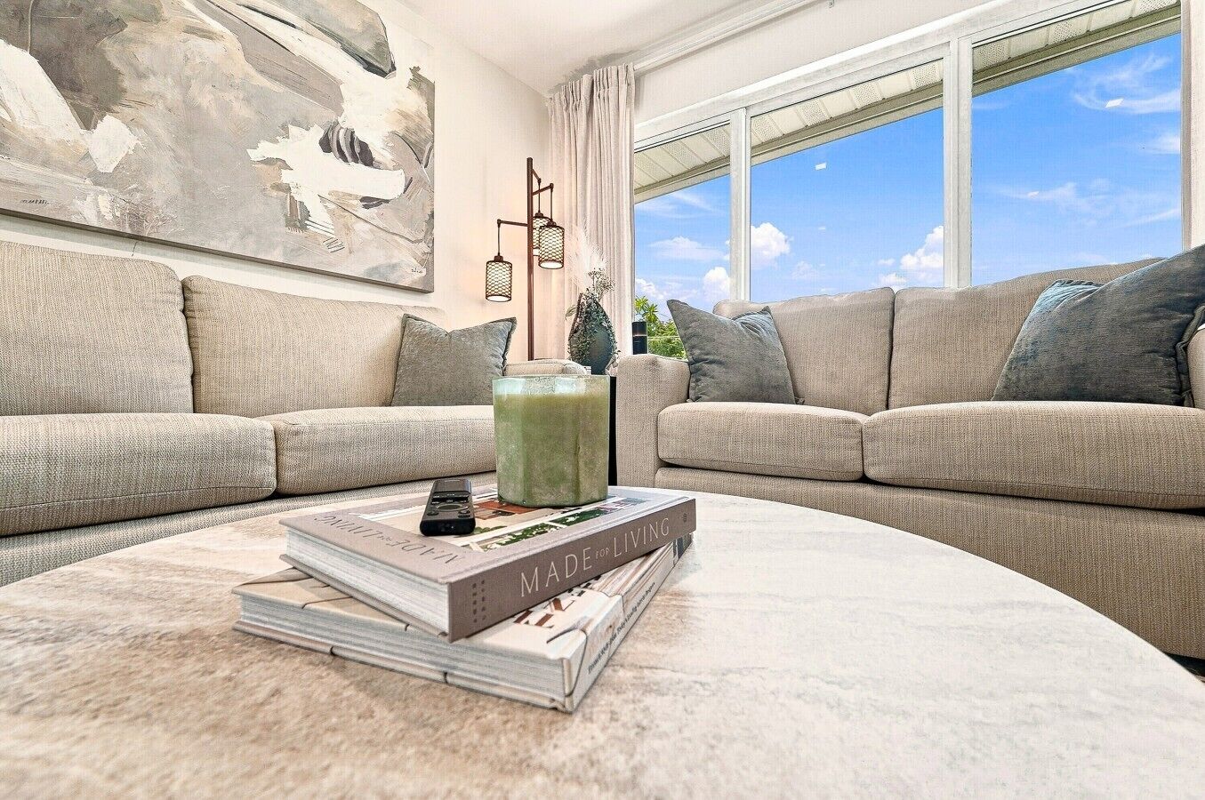 Un salon avec un canapé beige, une table basse sur laquelle sont posés des livres et une télécommande, un tableau au mur et une fenêtre donnant sur un paysage.