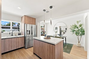 A modern kitchen with wooden cabinets, stainless steel appliances, a central island, and a dining area with a round mirror. - Spa Hot Tub | Mins to Beach | 2 Kings | Brand New! (Pompano Beach)
