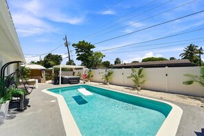 Ein Schwimmbecken mit Wohnbereich, weißer Liegefläche, weißem Zaun und überdachter Sitzgruppe mit Pergola.