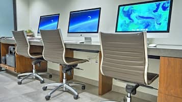A row of office chairs with wheels, a desk with a computer monitor, and a printer on a shelf.