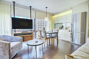 A modern living room with a flat-screen TV, a dining area with a table and chairs, a kitchen with a refrigerator, and a large window with curtains.