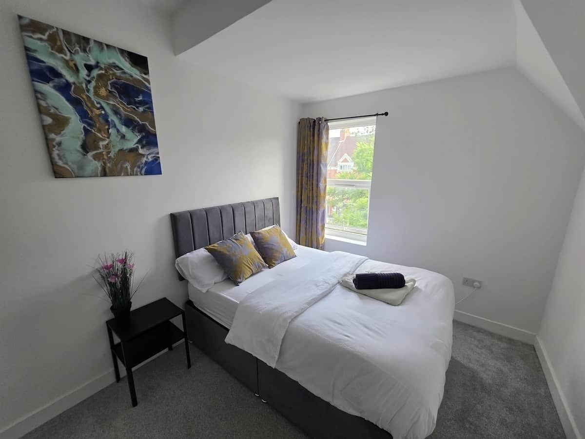 A bedroom with a bed, a nightstand, a painting, and a window with curtains.