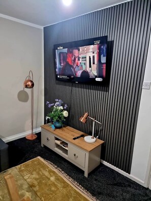 Un salon moderne avec un téléviseur à écran plat fixé au mur, une console en bois agrémentée d’objets décoratifs, une lampe sur pied et un tapis à motifs.