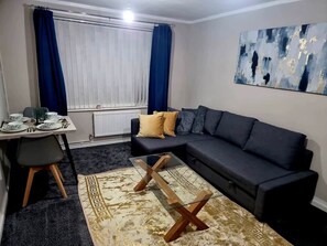 Ein Wohnzimmer mit einem dunklen Sofa, einem Glastisch, einem Esstisch mit Tellern und einem blauen Vorhang.