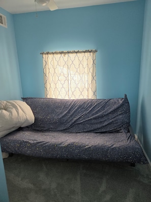 Ein Zimmer mit einem Einzelbett, einem Fenster mit gemusterten Vorhängen und einem Deckenventilator.
