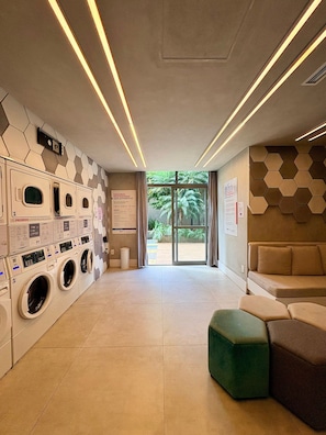 A modern laundry facility with multiple washing machines, a seating area with a green ottoman, and a view of the outdoors through a glass door. - Z Pinheiros  (São Paulo)