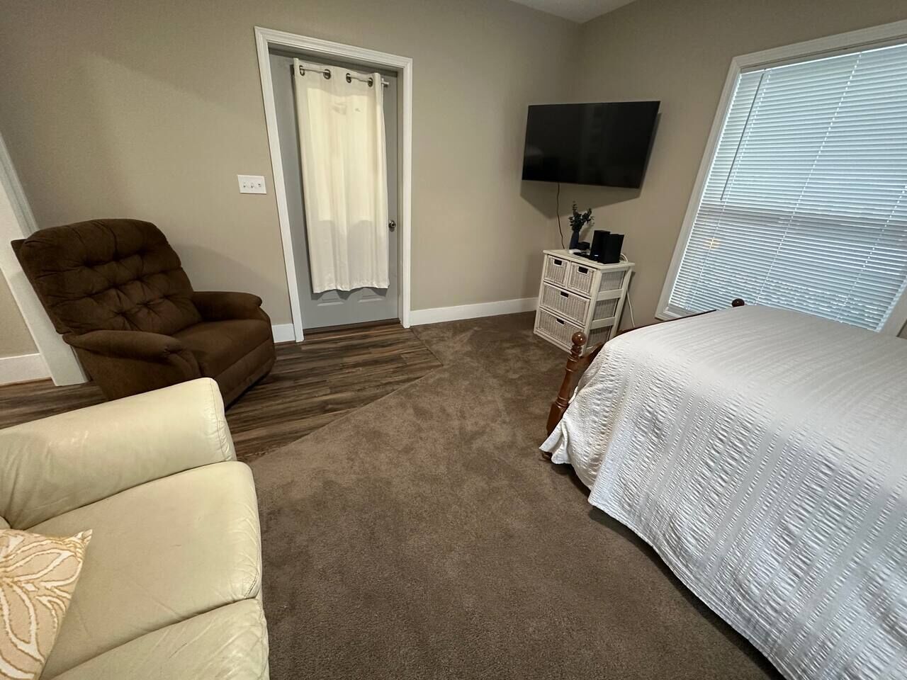 Une chambre à coucher avec un lit, un fauteuil marron, un fauteuil blanc, une télévision et une fenêtre avec des stores.