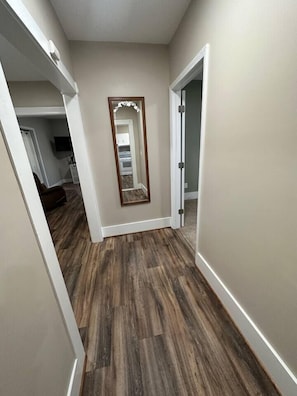 A hallway with wooden flooring, a mirror, and a door leading to another room.