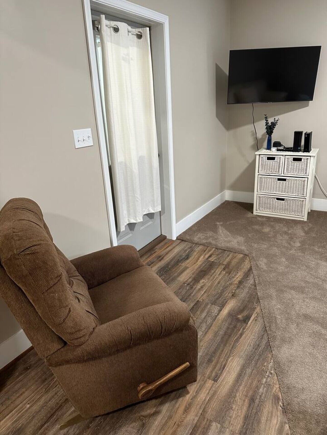Une pièce avec un fauteuil marron, un téléviseur fixé au mur et un meuble de rangement blanc.