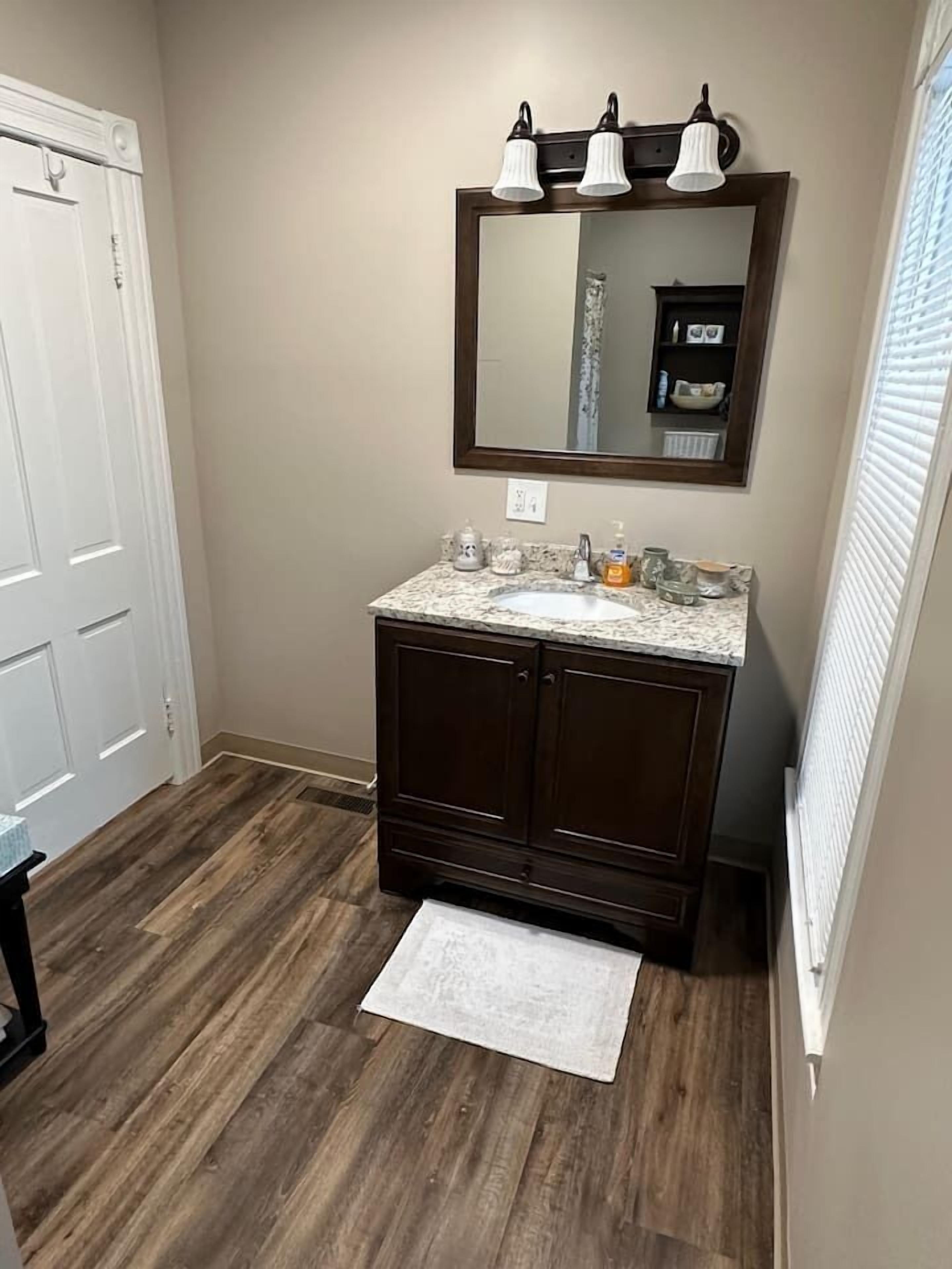 Baño con piso de madera, puerta blanca, tocador con espejo y ventana con persianas.