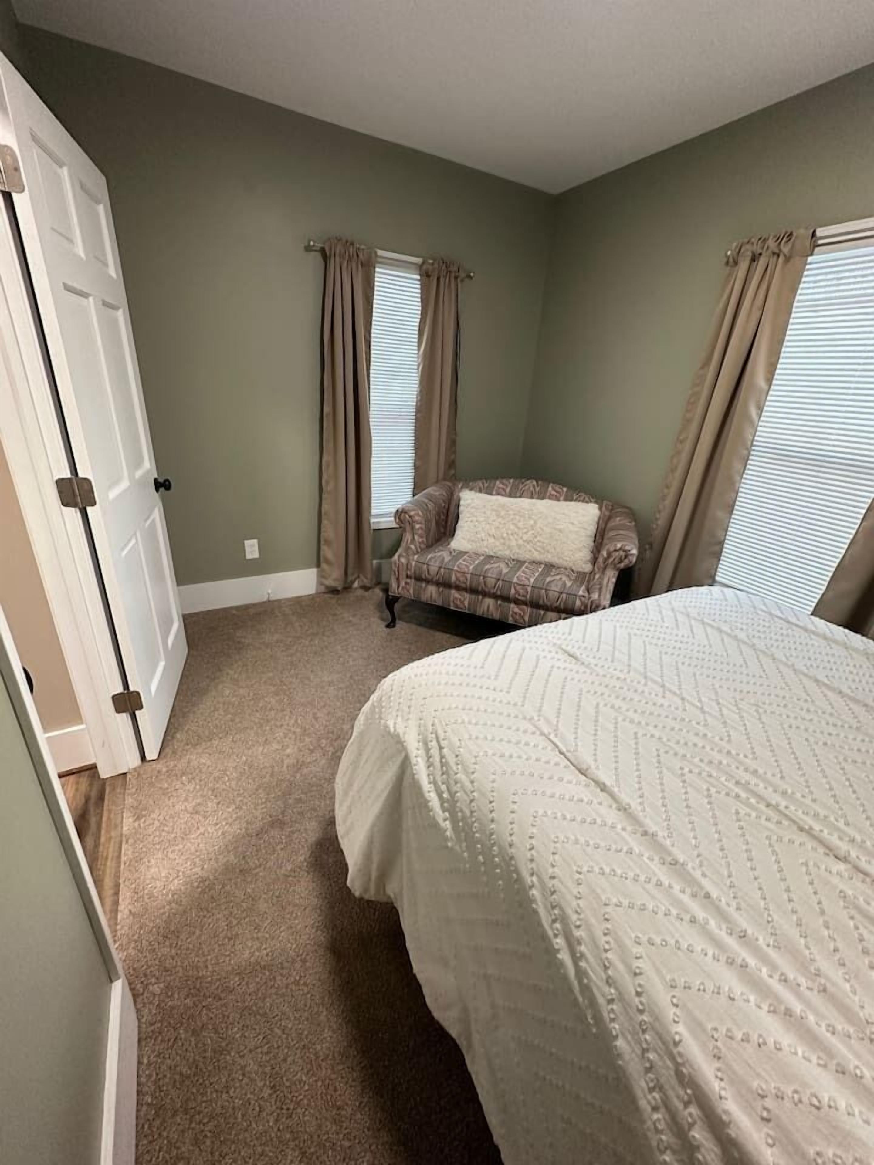 A bedroom with a bed, a chair, a door, and a window with curtains.