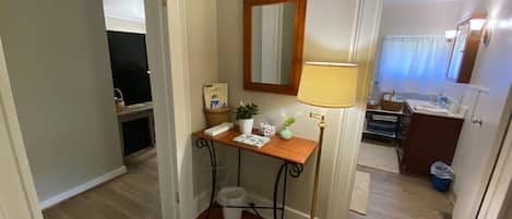 Un couloir avec un plancher en bois, un bureau avec une lampe, un miroir et une petite poubelle.