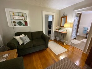 Una sala de estar con un sofá verde, piso de madera, alfombra blanca, una mesita y una estantería con objetos decorativos.