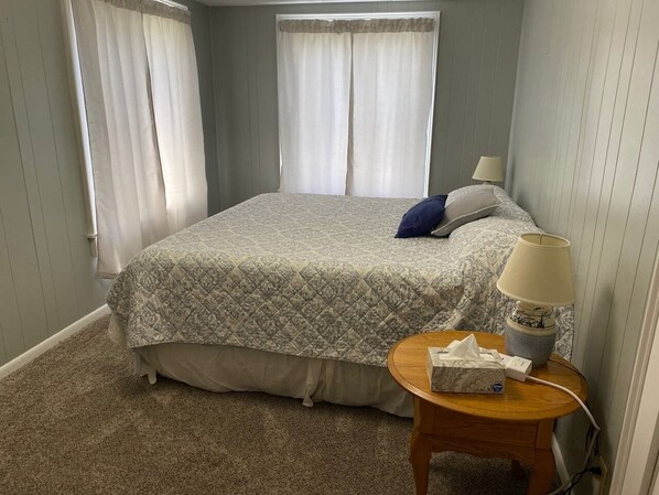 Un dormitorio con cama, cortinas, una lámpara de noche y una mesita de noche con un box de pañuelos.