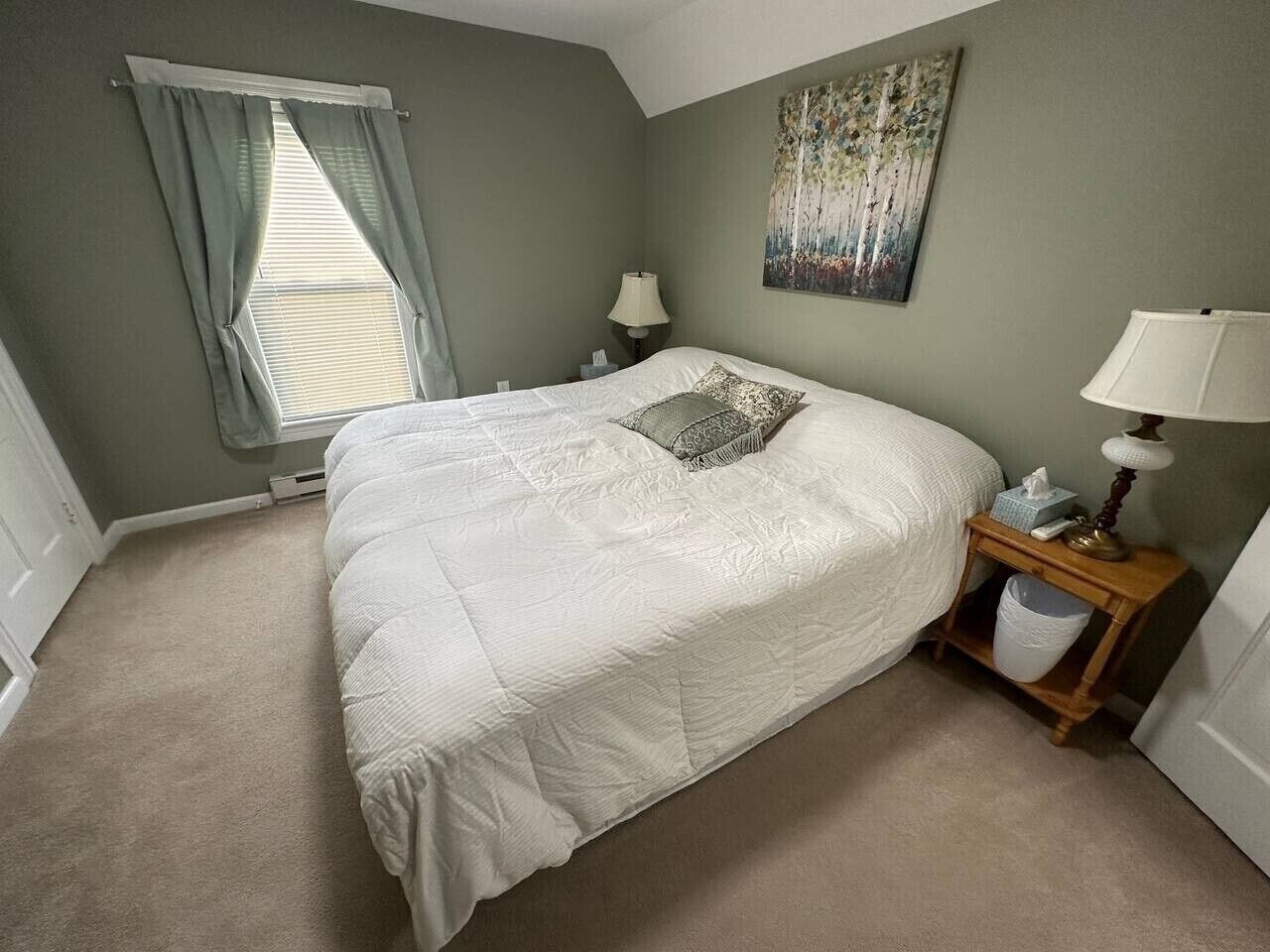 Une chambre à coucher avec un lit, une table de chevet, une lampe, une fenêtre avec des rideaux et un tableau au mur.