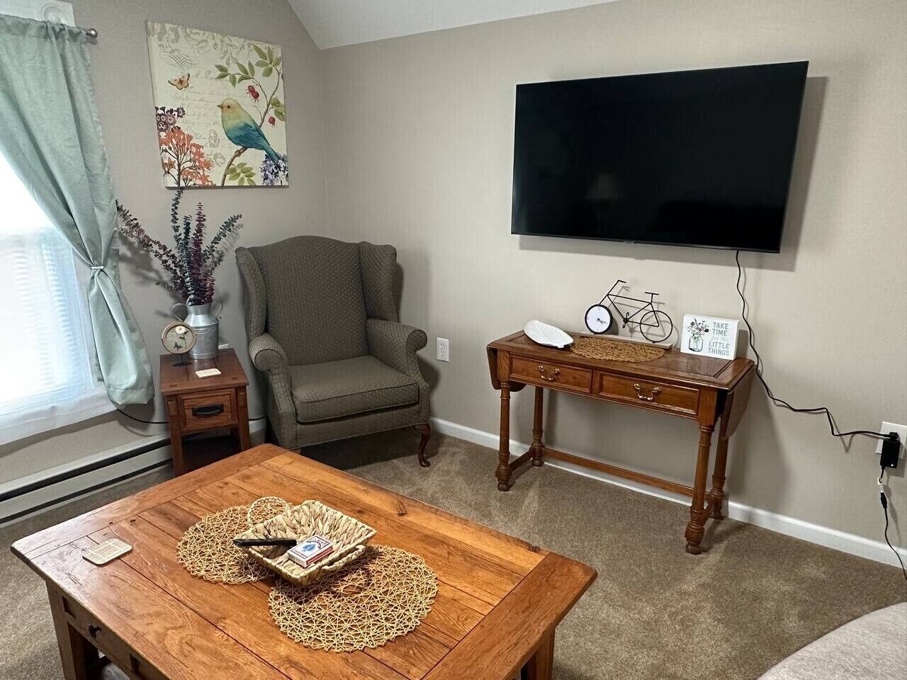 Un salon comprenant une table basse en bois, un canapé, un téléviseur fixé au mur, une petite table avec une horloge et des objets décoratifs, ainsi qu’une fenêtre avec des rideaux.