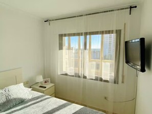 A hotel room with a bed, a bedside table with a lamp, a TV mounted on the wall, and a window with sheer curtains. - Apartment in Salou, 2 minutes from the beach (Salou)