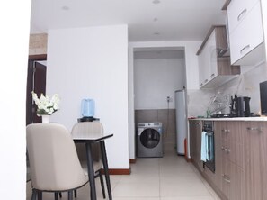 A compact kitchen with a washing machine, a dining area with a chair, and a small table. - Imagine Luxury- 2 Bedroom (Nairobi)