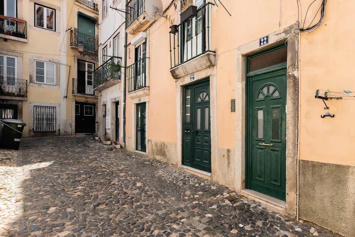 Une ruelle étroite pavée, bordée d’immeubles aux portes et balcons verts.