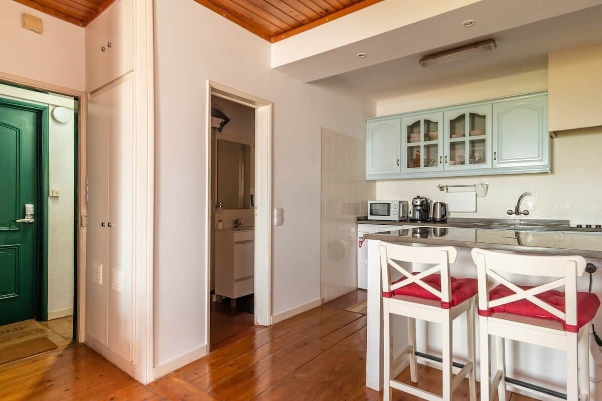 A kitchen with white cabinets, a microwave, and a coffee maker. There are two white bar stools with red cushions.