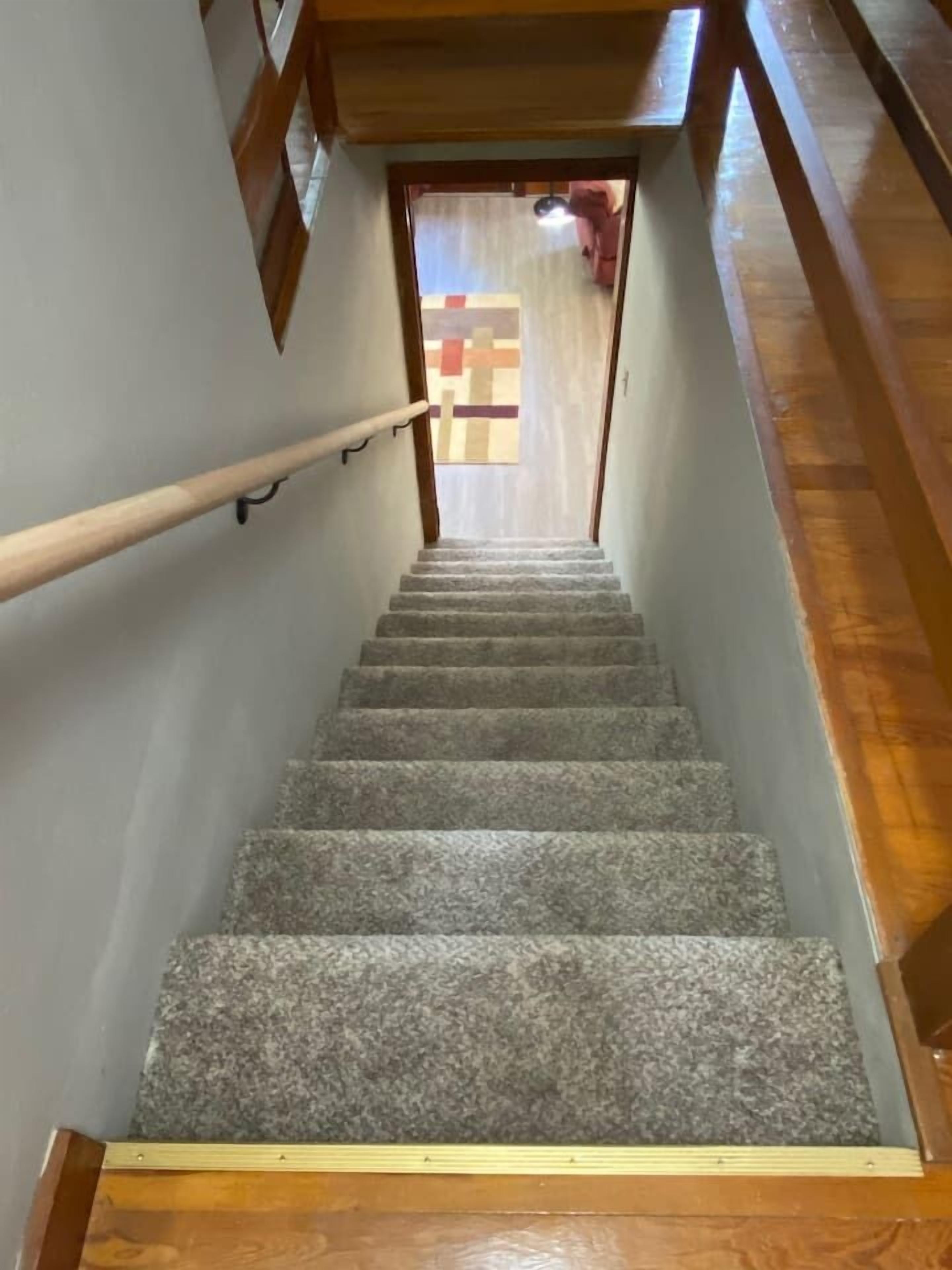 Un escalier avec des rampes en bois et des marches recouvertes de moquette, menant à une porte ornée d’un motif géométrique.