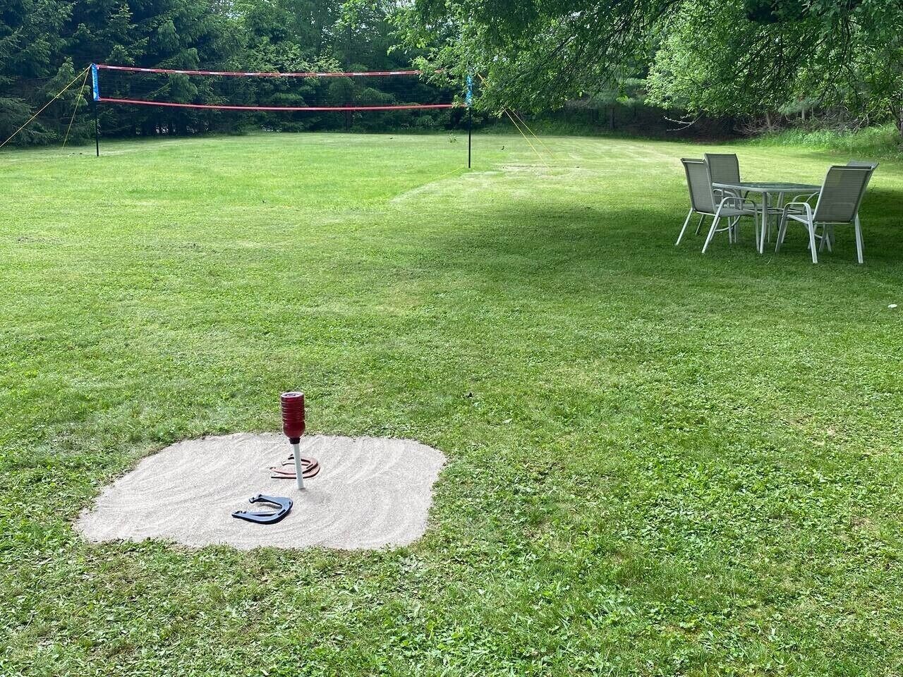 Un espace extérieur verdoyant avec un filet de volley-ball, une table de pique-nique avec des chaises et une couverture sur laquelle se trouvent une paire de tongs et un objet rouge.