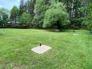 A grassy outdoor area with a circular patch of dirt, a metal pole, and a netted structure in the background. - Rushford Lakeside Family Getaway (Houghton)