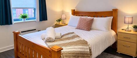 Una cama bien hecha con cabecera de madera, una mesita de noche con lámpara, una pequeña planta en maceta y una ventana con cortinas.