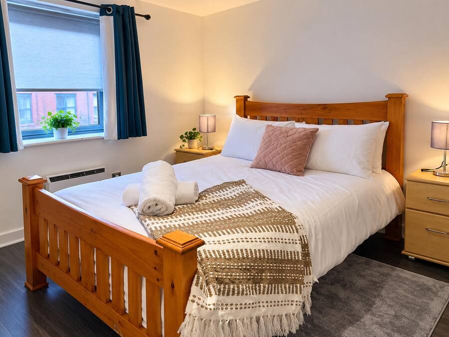 A neatly made bed with a wooden headboard, a bedside table with a lamp, a small potted plant, and a window with curtains.