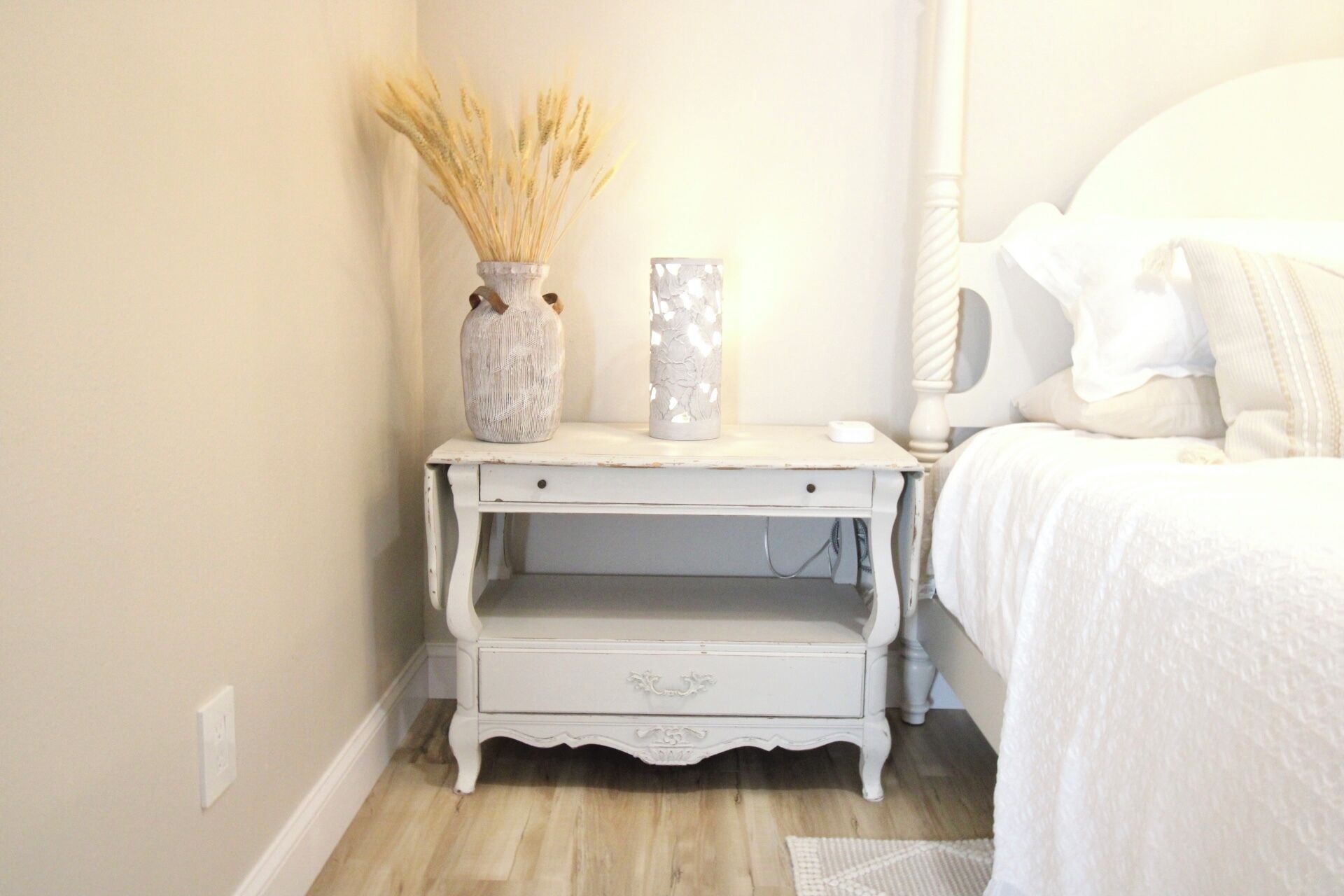 A white bedside table with a vase and a candle, a drawer, and a bed with white bedding.