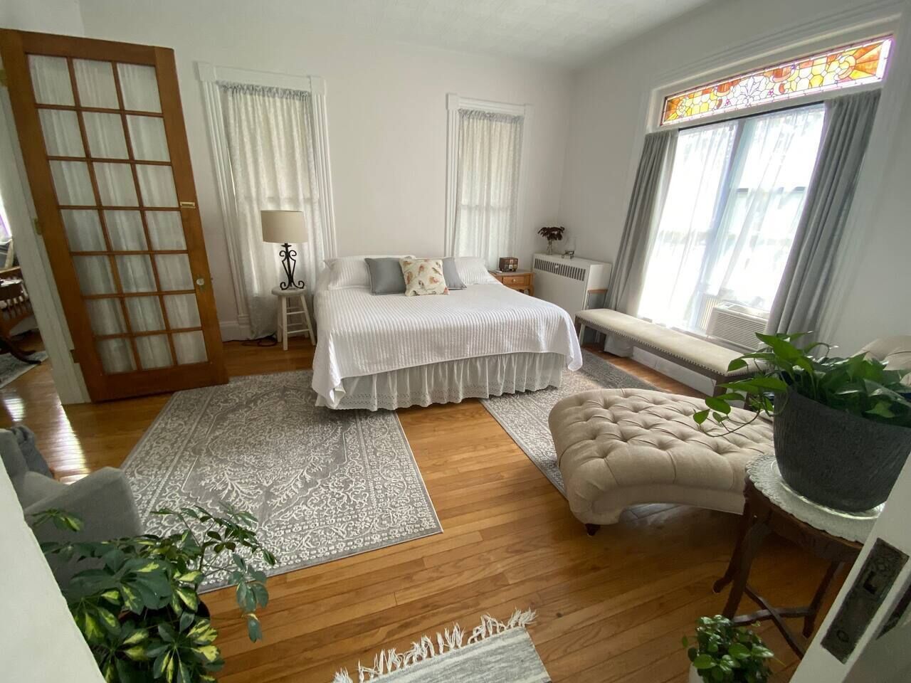 Une chambre à coucher avec un lit, des tables de chevet, une fenêtre avec des rideaux, un radiateur, une plante en pot et un plancher en bois.